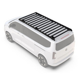 A black Front Runner Volkswagen T7 Transporter Caravelle LWB (2025-Current) Slimsport Roof Rack Kit with crossbars mounted on the roof of a white van, shown from a high-angle perspective on a clean white background.