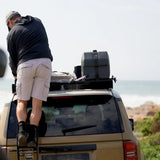 A man in a black hoodie and khaki shorts climbs onto the back of a tan Land Rover Prado SUV, loading gear onto the roof rack near the ocean the Front Runner Pro Water Tank With Mounting System / 42L is fitted to the rack.