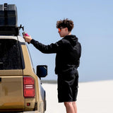 A man in a black hoodie and black cargo shorts stands next to a tan SUV, filling a pink water bottle from the Front Runner Pro Water Tank With Mounting System / 42L on the vehicle's roof rack. The background is a clear blue sky and white sand dunes.