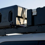 Close-up of a rooftop-mounted Front Runner Pro Water Tank With Mounting System / 42L with a hose connection and a visible Front Runner logo, installed on a vehicle roof against a pale sky.