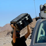 Person carrying a Front Runner Wolf Pack Pro Ammo Box over their head in a desert setting with a vehicle and mountains in the background.