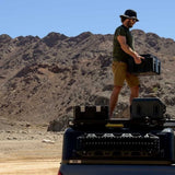 Person standing on a Camp 4x4 Rentals vehicle with a Front Runner Wolf Pack Pro Hi-Lid Ammo Box in a desert landscape