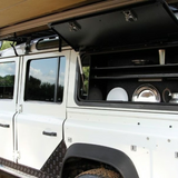 A white Land Rover utility vehicle with its Front Runner Land Rover Defender (1983-2016) Gullwing Window side cargo/storage compartment open, showing shelves and silver cookware inside. The vehicle is photographed outdoors with trees and sky visible in the windows.