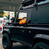 A black off-road vehicle with a roof rack and Front Runner Land Rover Defender (1983-2016) Gullwing Window, parked at a gas station. A person wearing a bright orange jacket is seated in the driver's seat, visible through the open window.