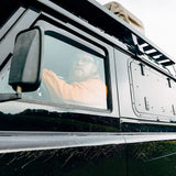 A man wearing glasses and an orange shirt sits inside a black vehicle, visible through the side window. The photo is taken from a low angle, focusing on the driver's area and the side of the vehicle with a Front Runner Land Rover Defender (1983-2016) Gullwing Window.