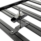 Close-up of a Front Runner Series Spare Wheel Clamp, black metal roof rack accessory with a wing nut adjustment bolt and mounting bracket attached above parallel roof rails on a white background.