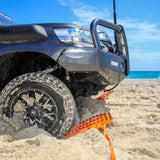 An orange MAXTRAX XTREME traction mat with a black handle is deployed from under a dark gray off-road vehicle on a sandy beach with the ocean and blue sky in the background.