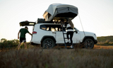 Two people interacting with a white SUV equipped with a Front Runner Feather-Lite II Rooftop Tent in a natural setting.