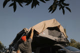 Person setting up a Front Runner rooftop tent on a car with a clear sky and trees in the background