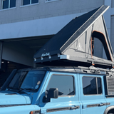Blue vehicle with a rooftop tent and Alu-Cab INEOS Load Bar Risers attached in front of a building