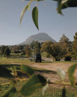 Suzuki Jimny on a dirt road with a mountain in the background, framed by green leaves.