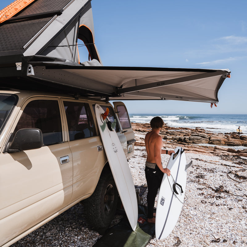 Person with surfboards standing next to a Camper with an extended Alu-Cab 270 Shadow Awning and a Gen3.R rooftop tent on a beach. Camp4x4. camp 4x4, overlanding, camping. Double Cab. Single Cab. Extra Cab