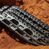 Black MAXTRAX LITE with aggressive tread knobs, partially embedded in reddish-brown dirt.