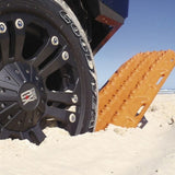 An Orange MaxTrax MKII with aggressive treaded spikes, shown partially deployed from a vehicle on a sandy beach.