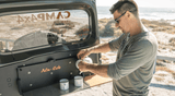 A man pouring coffee into camping mugs on an Alu-Cab shelf at the back of the Suzuki Jimny 4x4 on the beach front, available for rent.