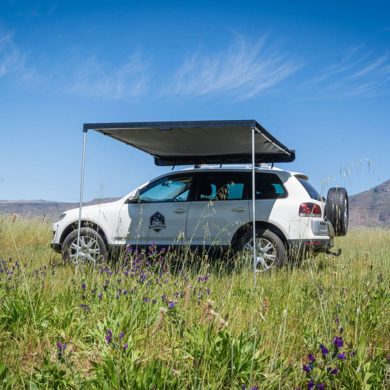 White SUV with the RoofPod Awning out, in a grassy field with mountains in the background
