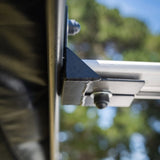 Close-up showing the mounting bracket of the RoofPod awning on a vehicle with blurred greenery in the background
