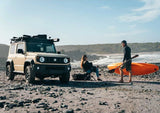 Two people by a beach with a Suzuki Jimny 3 door fitted with a Front Runner Suzuki Jimny Slimline II Roof Rack, and an orange kayak.