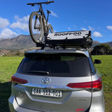 Silver SUV with a RoofPod SoloPod rooftop tent and bicycle on a green field