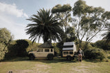 Suzuki Jimny 4x4 with rooftop tent and fully kitted trailer parked in a grassy area with trees and a palm tree in the background.