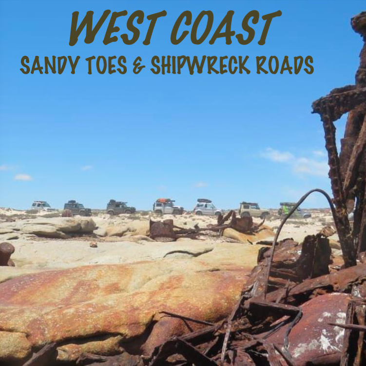 Rusted shipwreck on a rocky beach with 4x4 vehicles in the background under a clear blue sky.