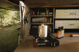 Portable stove with a pot on top inside a vehicle, with camping supplies around.