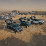 Three off-road pickup trucks parked on a desert canyon landscape, each equipped with an iKamper rooftop tent and ladder access, photographed from above at sunset.