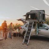 A Hardshell iKAMPER Skycamp 4.0 Trio, 3 sleeper, rooftop tent mounted on the roof of a silver SUV at a campsite during sunset, with a ladder extending from the side and two people preparing food near a small table nearby.