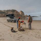 A family and two dogs camp on a sandy beach with a dark gray SUV featuring a Hardshell iKAMPER Skycamp 4.0 Trio, 3 sleeper, rooftop tent and ladder. A small table holds camping gear, with the group standing nearby against a backdrop of rocky cliffs and the ocean.