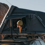 A person wearing a tan beanie sits inside a black IKAMPER rooftop tent mounted on a vehicle roof rack, with rocky mountains and a clear sky in the background.