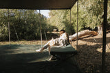 Person lying on a stretcher mattress under a canopy in a natural setting with trees and ground cover.